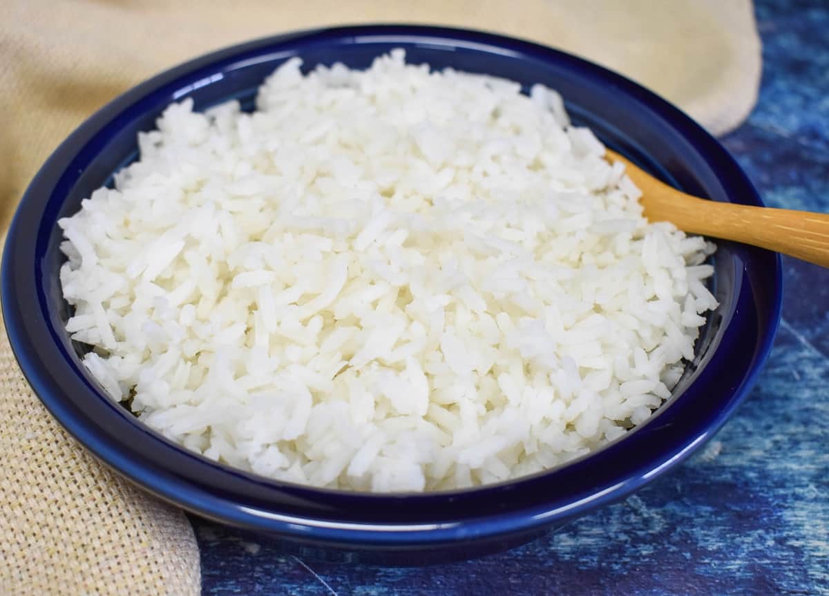 A close up of rice served in a blue bowl with a wooden spoon displayed on a blue table with a beige linen.