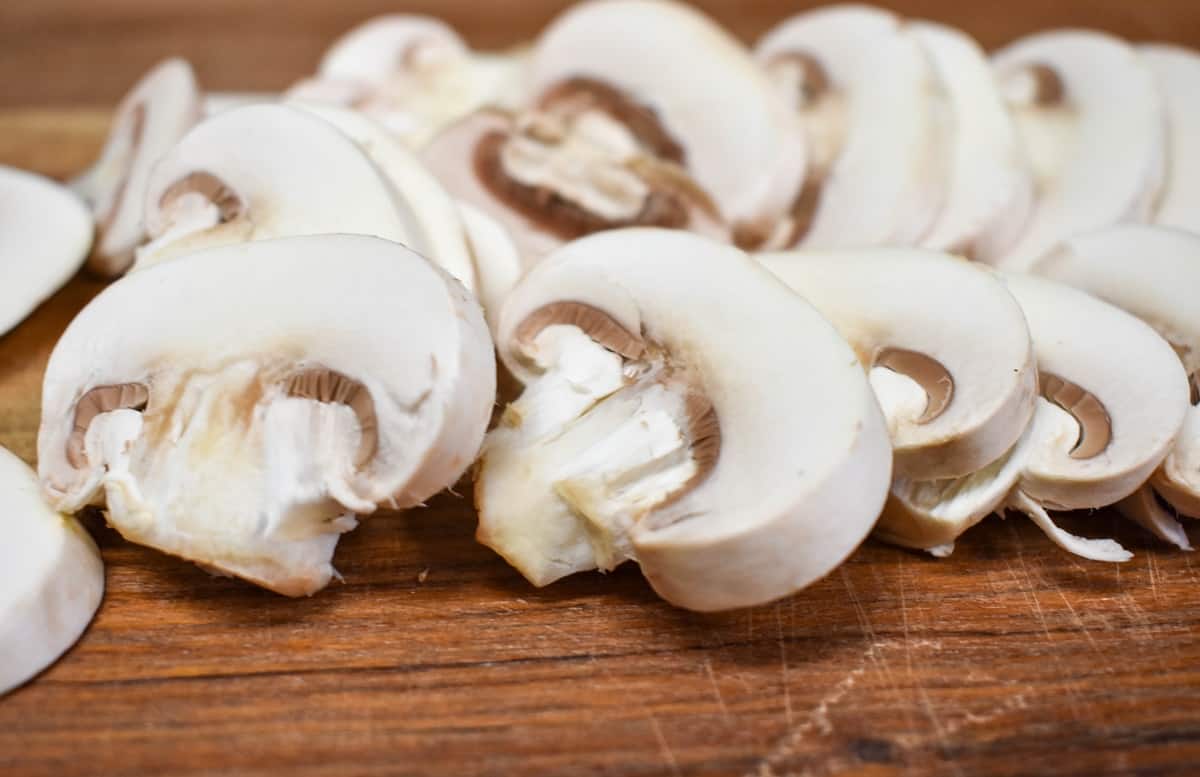 A close-up image of sliced white button mushrooms on a wood cutting board.