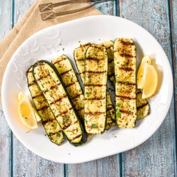 Grilled zucchini arranged on a white platter, garnished with chopped parsley and served with lemon wedges.