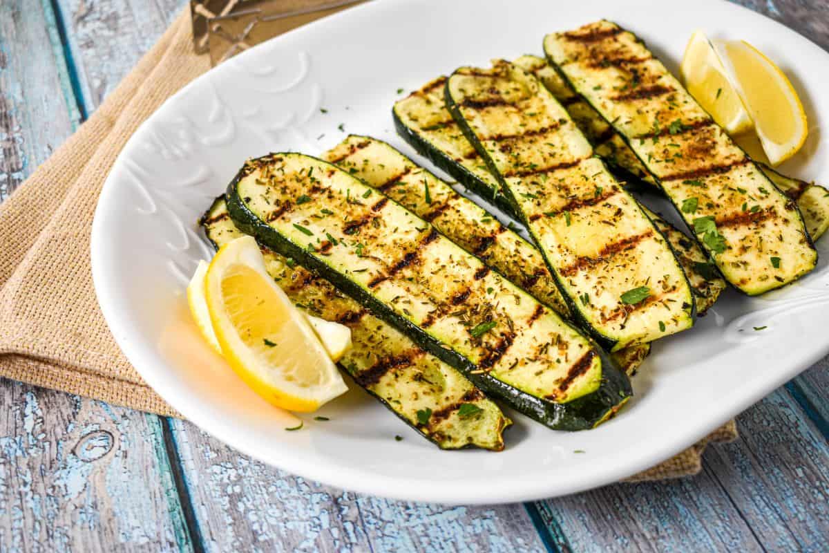 Grilled zucchini arranged on a white platter, garnished with chopped parsley and served with lemon wedges.