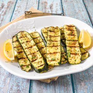 Grilled zucchini arranged on a white platter, garnished with chopped parsley and served with lemon wedges.
