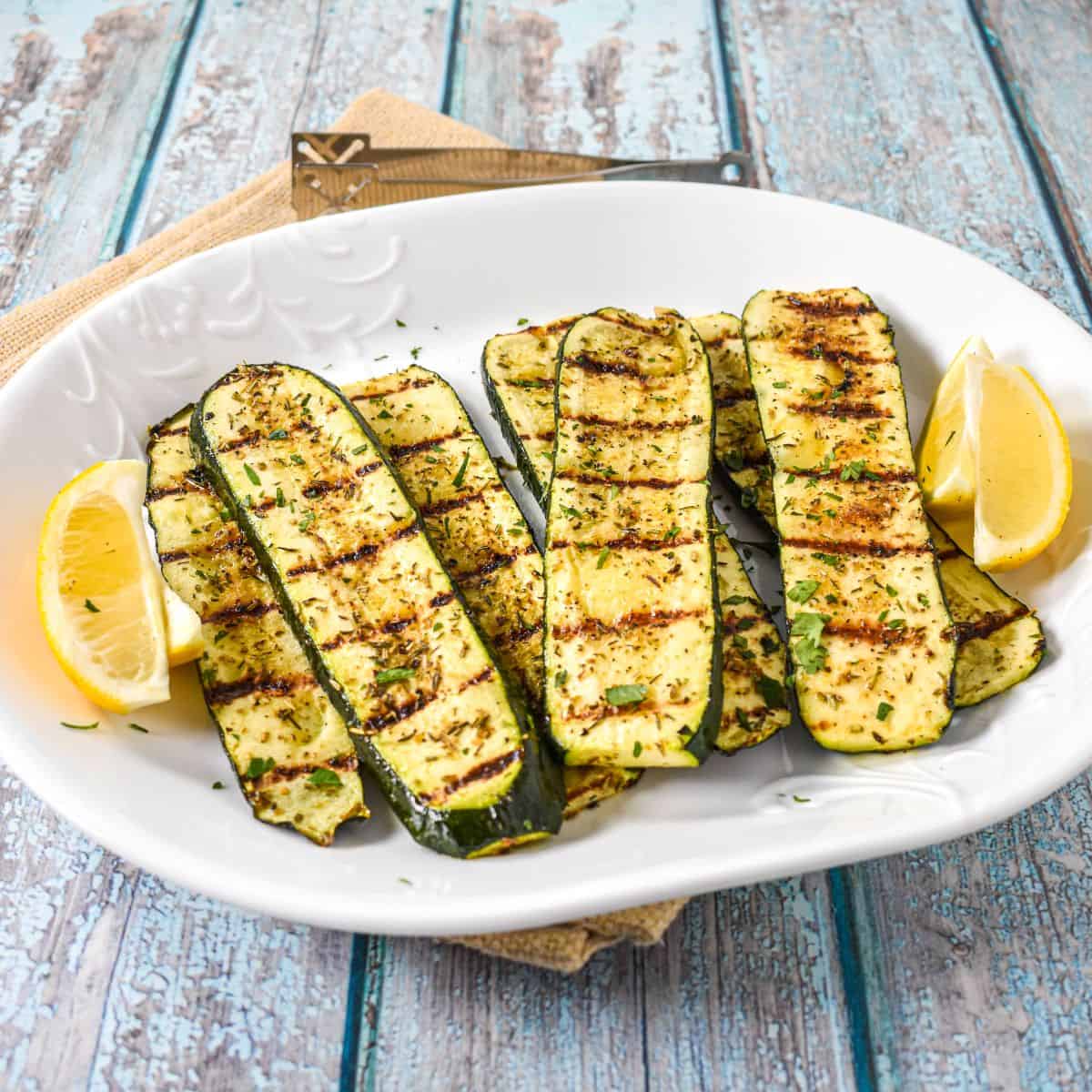 Grilled zucchini arranged on a white platter, garnished with chopped parsley and served with lemon wedges.
