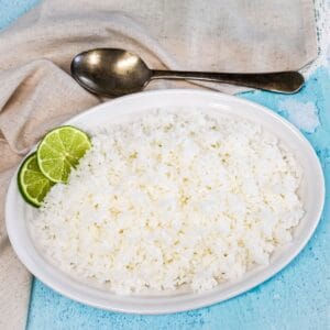 Cooked white rice served on a white platter, garnished with lime slices with a linen and serving spoon in the background.