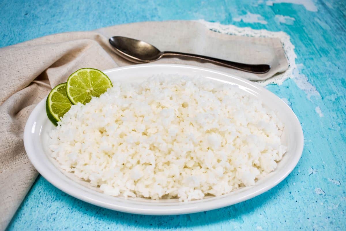 Cooked white rice served on a white platter, garnished with lime slices with a linen and serving spoon in the background.