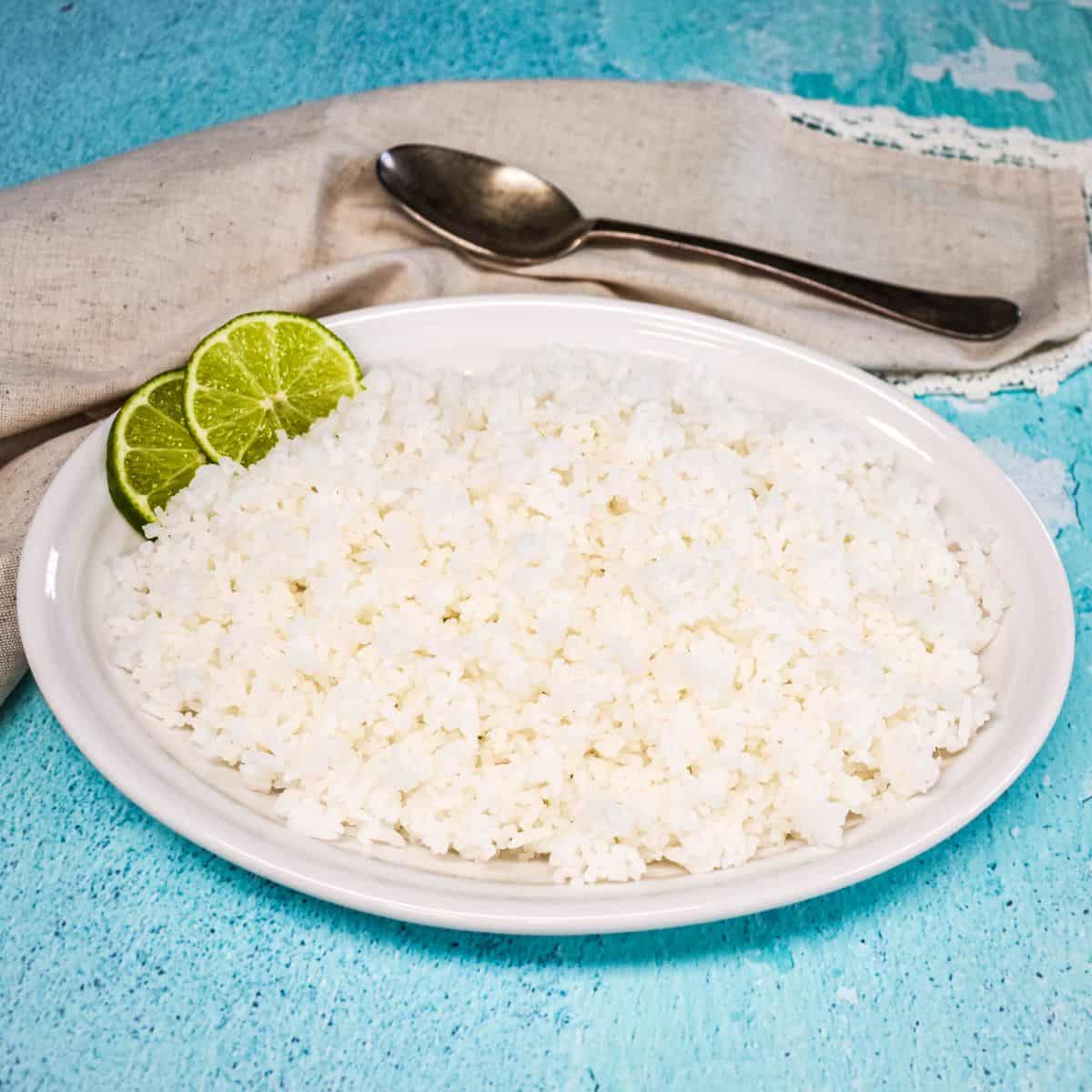 Cooked white rice served on a white platter, garnished with lime slices with a linen and serving spoon in the background.