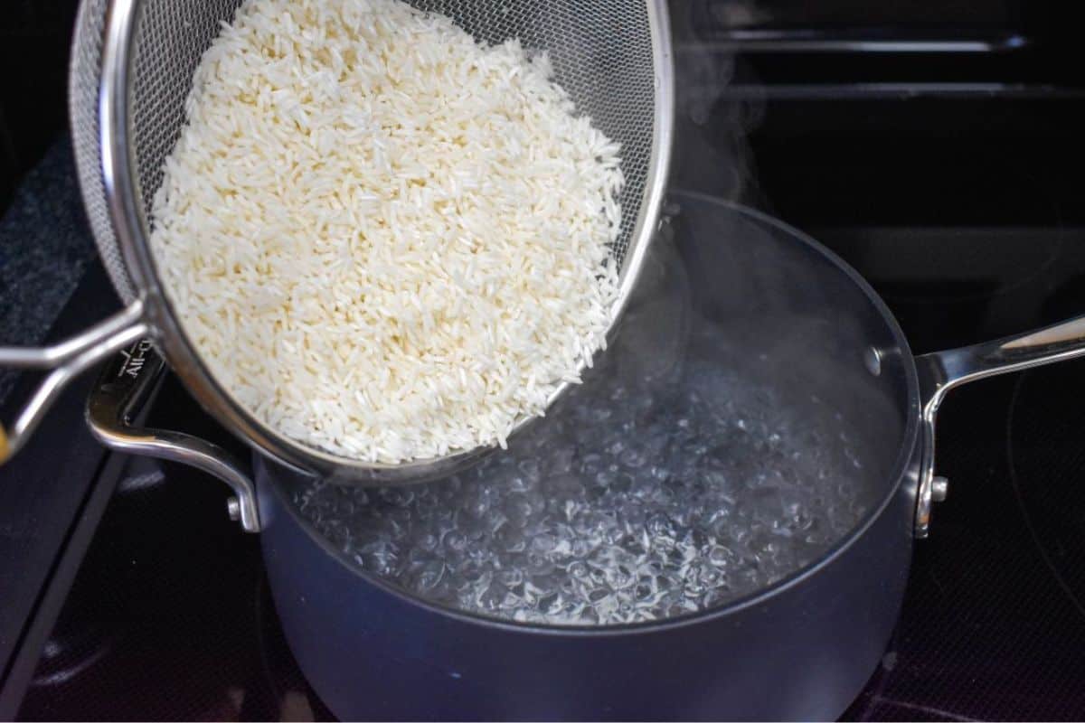 Rice in a mesh strainer being poured into water boiling in a black saucepan.