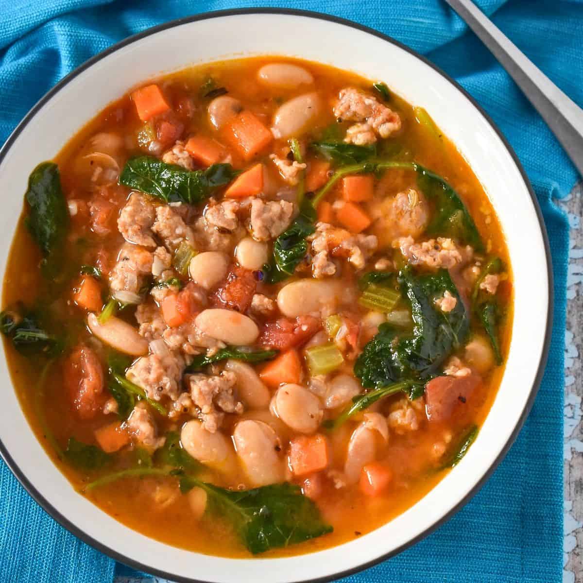 The sausage white bean kale soup served in a white bowl set on a blue linen.