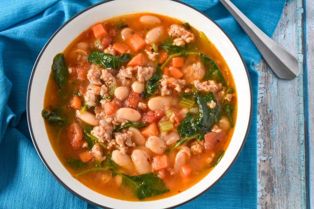 The sausage white bean kale soup served in a white bowl set on a blue linen.