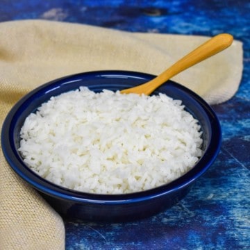 White rice served in a blue bowl displayed with a beige linen and a wooden spoon on a blue table.