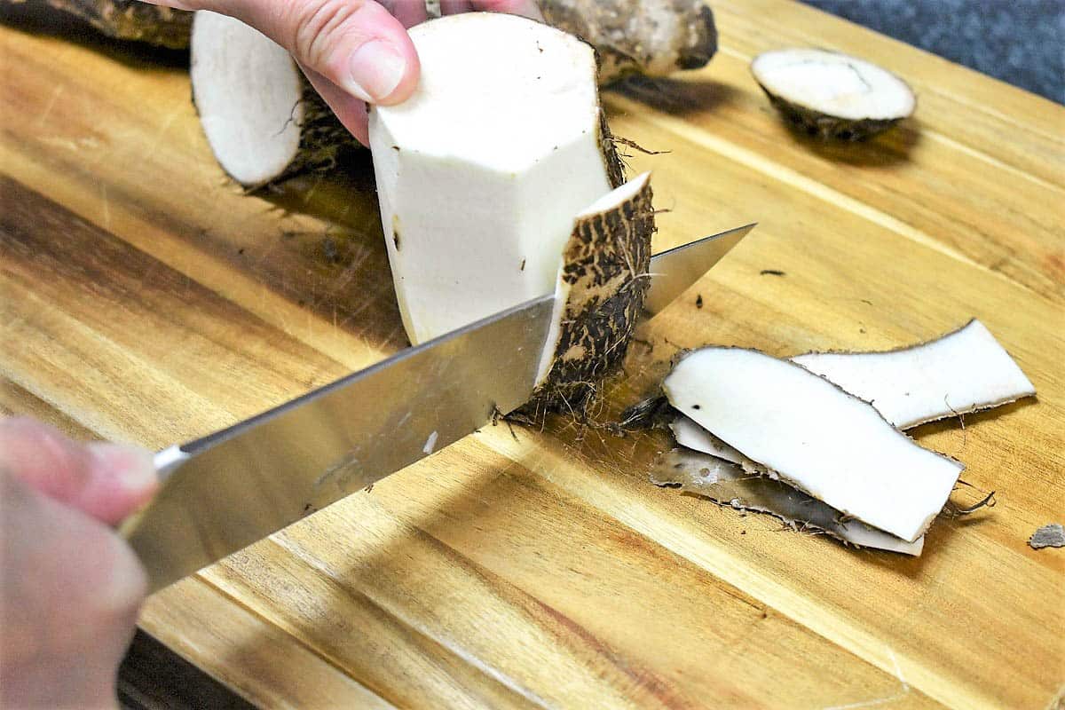 An image of a hand holding and peeling a malanga with a large knife on a wood cutting board.