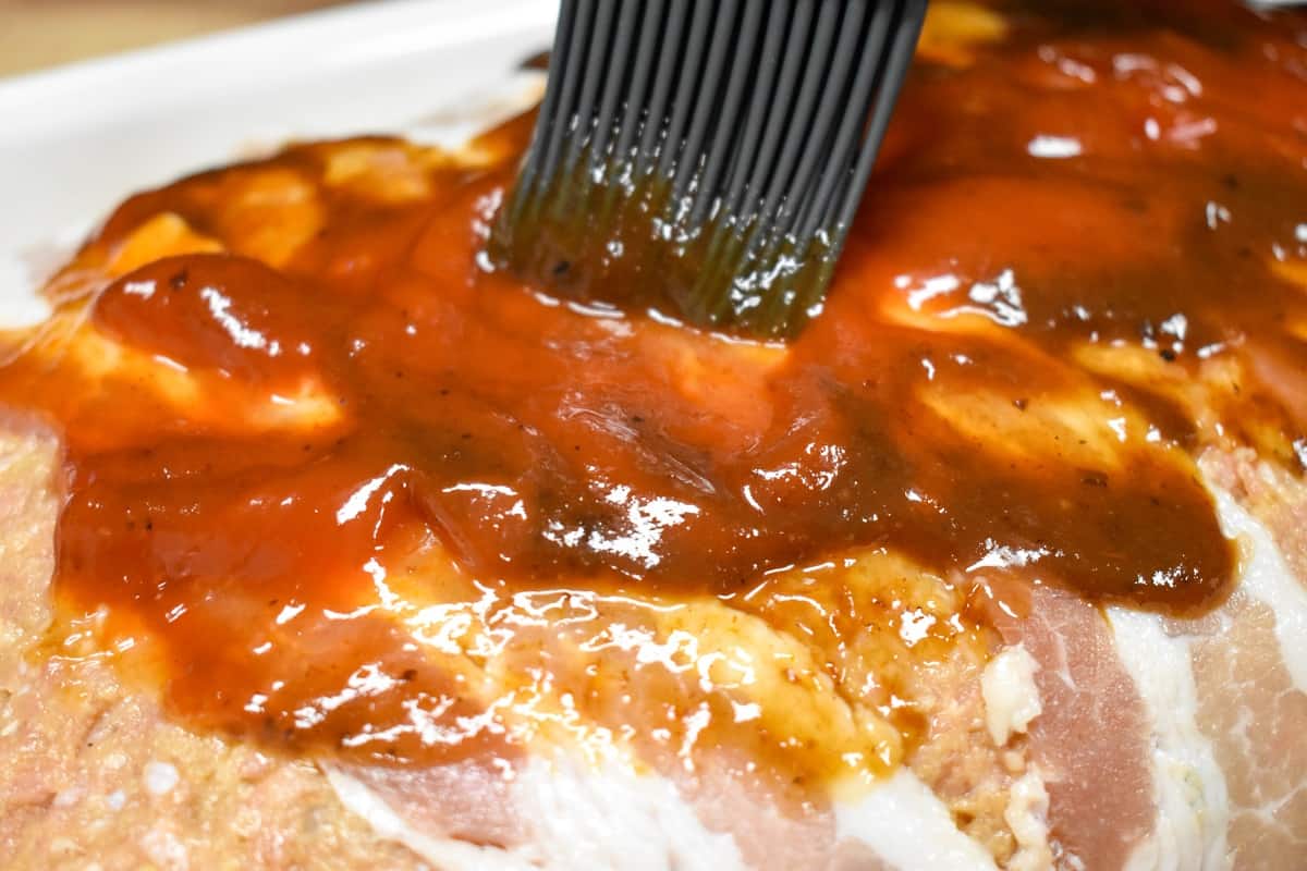 A gray silicone brush spreading a ketchup and steak sauce mixture on top of the uncooked meatloaf.