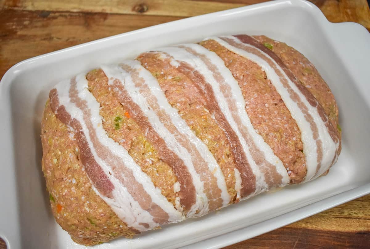Four bacon slices wrapped over the loaf in a white baking dish.
