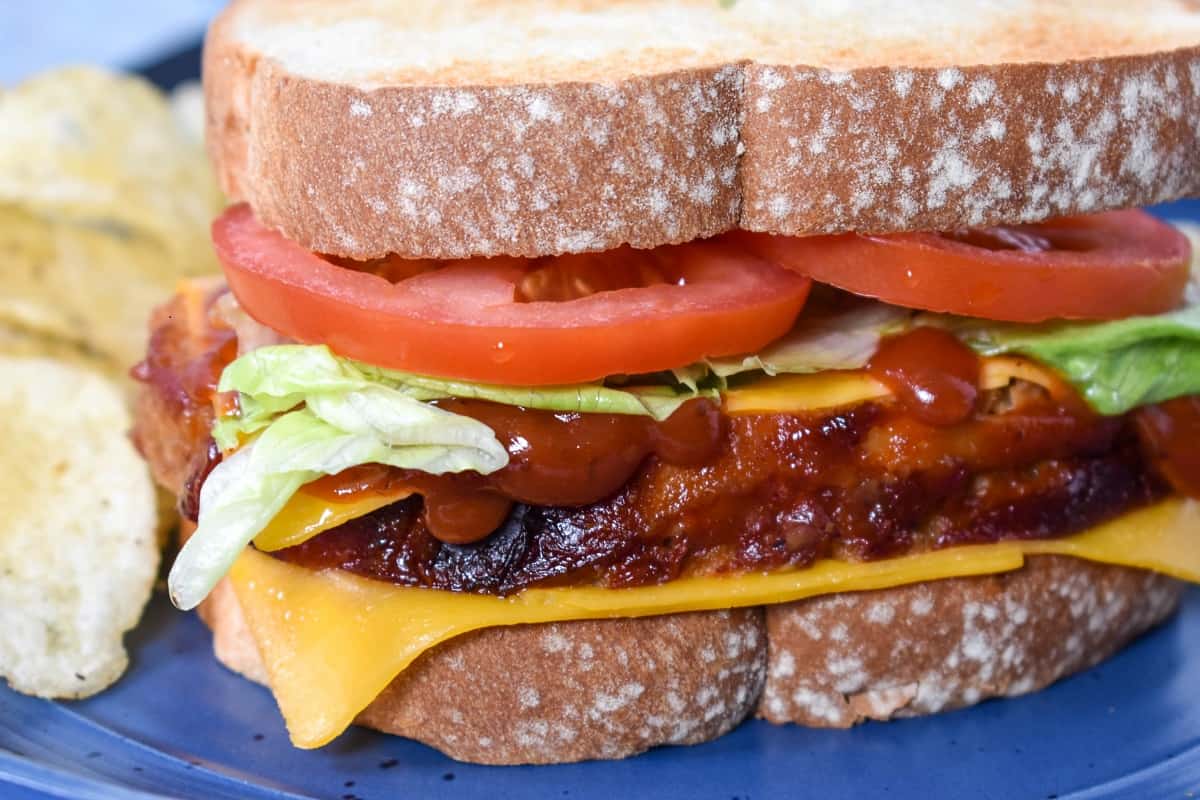 A close up image of the sandwich with cheddar cheese, meatloaf, cheddar again, sauce, lettuce and two tomato slices. It's served on a blue plate with chips in the background.
