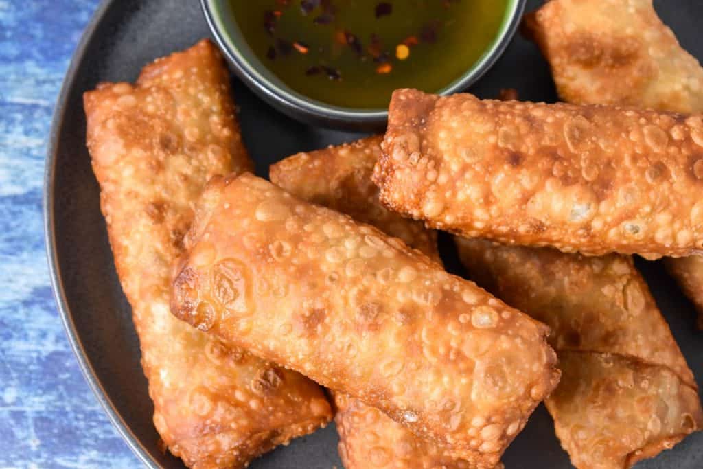 Six fried egg rolls arranged on a gray plate with a small bowl of sauce on the side.