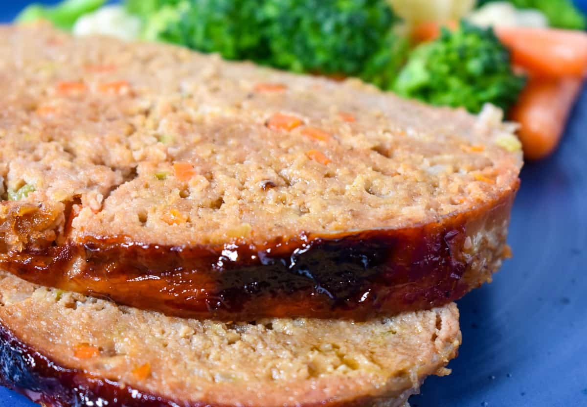 Two slices of meatloaf served on a blue plate with steamed broccoli, carrots and cauliflower in the background.