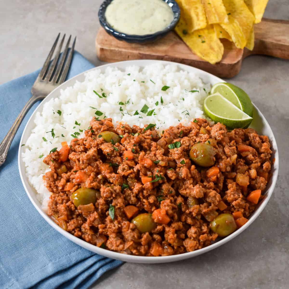 A white plate with ground turkey picadillo and white rice, garnished with chopped parsley and lime wedges, served with plantain chips and dipping sauce.