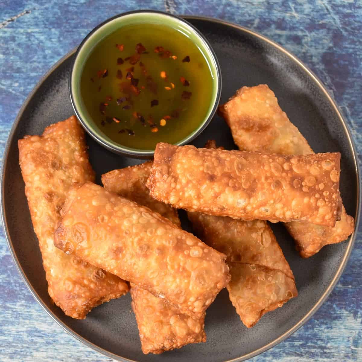Six fried egg rolls arranged on a gray plate with a small bowl of sauce on the side.