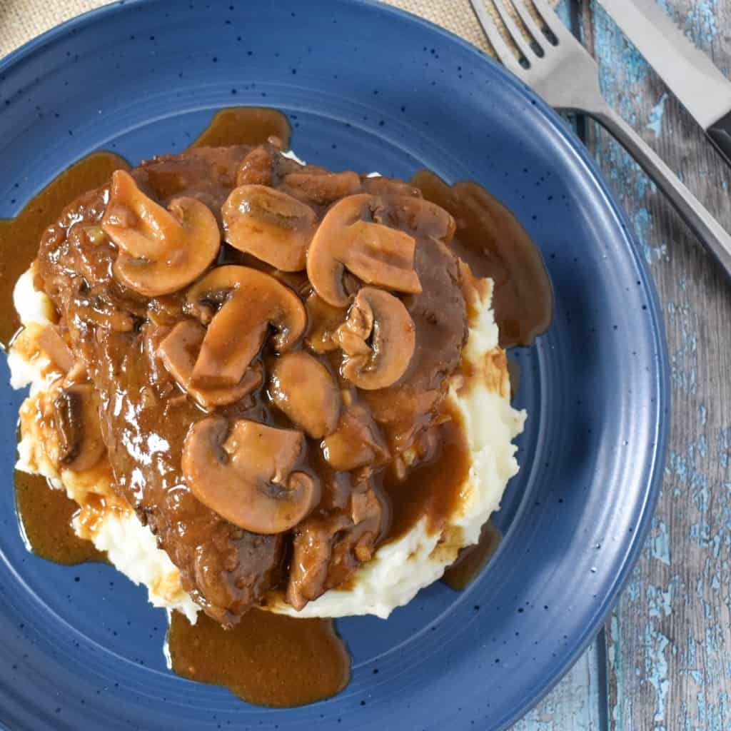 A thick sirloin steak covered in a dark brown mushroom gravy on a bed of mashed potatoes served on a blue plate.
