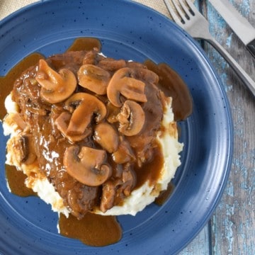 A thick sirloin steak covered in a dark brown mushroom gravy on a bed of mashed potatoes served on a blue plate.