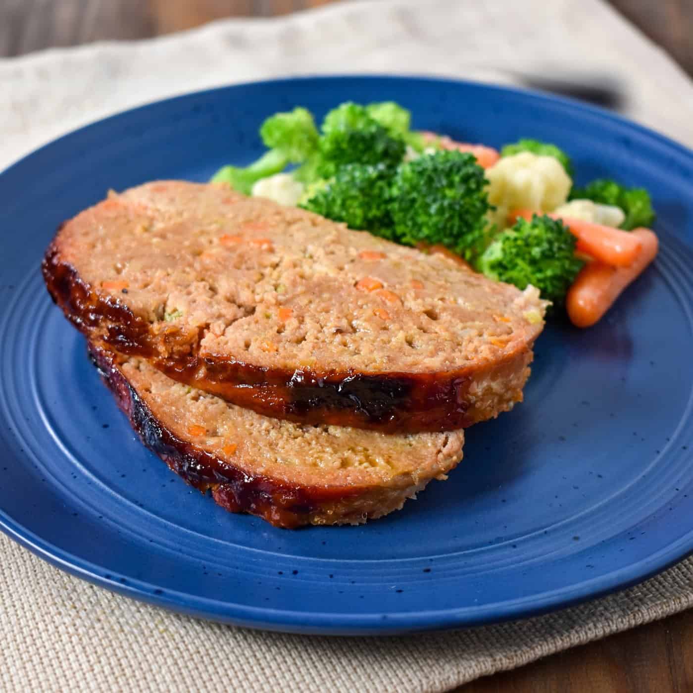Two slices of glazed turkey meatloaf on a blue plate with steamed broccoli, cauliflower, and baby carrots.
