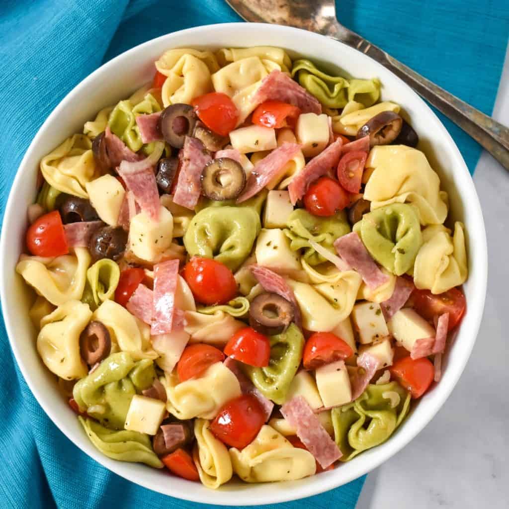 The tortellini pasta salad served in a large white bowl, displayed on a white table with an aqua colored linen underneath and a serving spoon in the background.