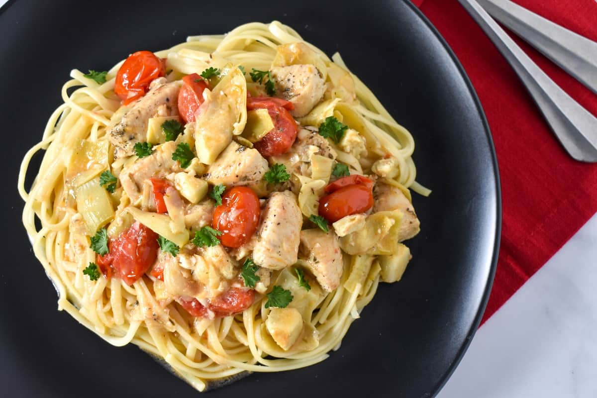 The artichoke chicken served on a bed of linguine noodles on a black plate set on a red linen.