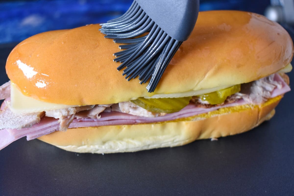 A gray silicone brush applying butter on the top slice of medianoche bread.