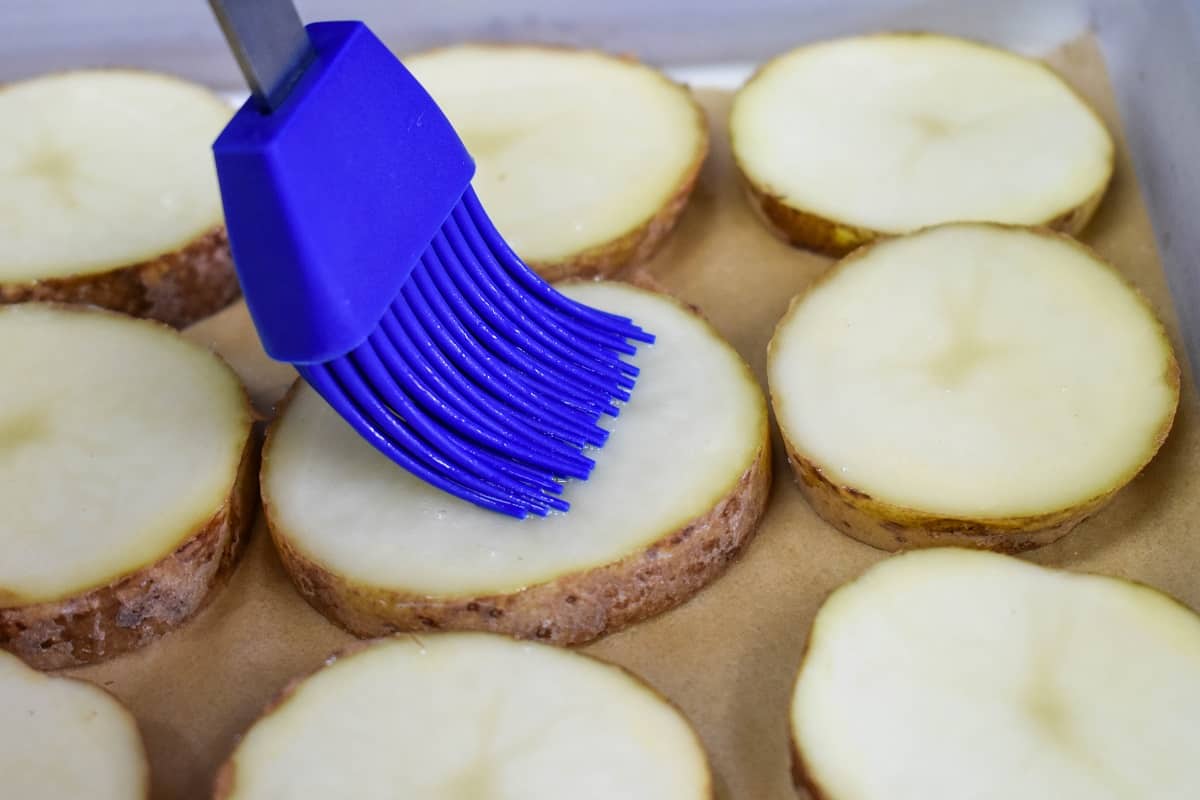 A blue silicone pastry brush applying olive oil on a potato slice.