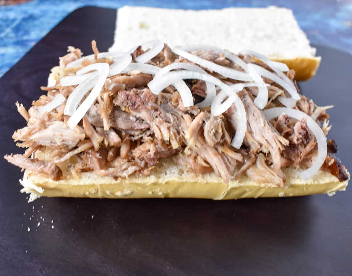 A generous amount of shredded roast pork topped with thin sliced onions on the bottom half of Cuban bread. The top part is in the background.