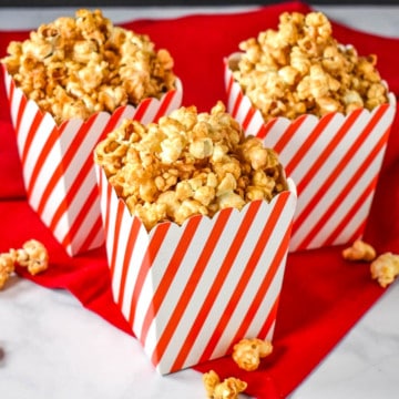 Three red and white striped containers filled with the caramel corn displayed on a red linen on a white table.