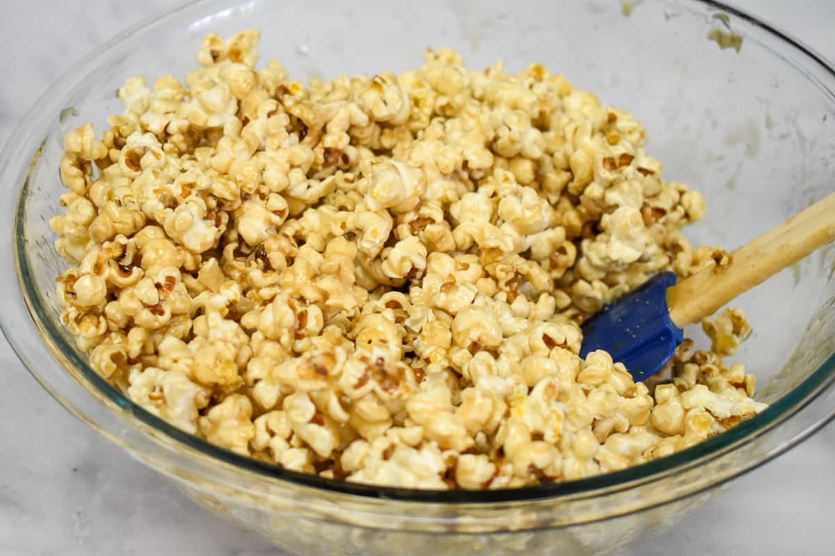 The popcorn coated with the caramel in a large, glass bowl with a blue silicone spatula on the right.