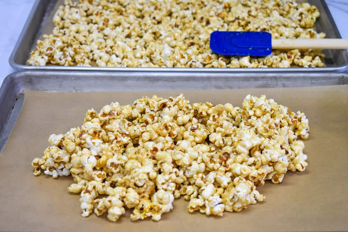 Two baking sheets the one in the back has the popcorn spread on it and has a blue spatula on it, the one in the front is not spread yet.