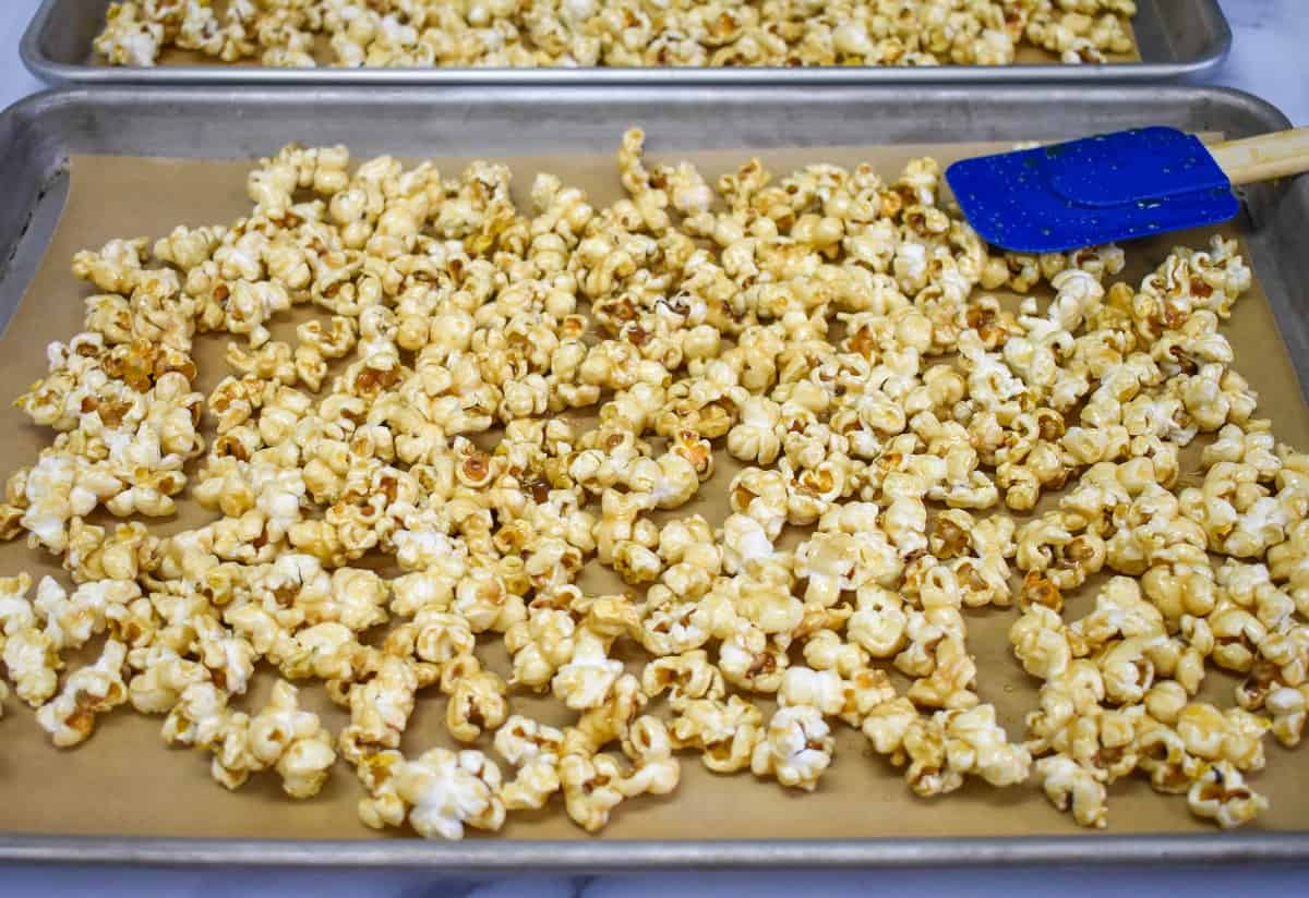 An image of the caramel corn spread on a large baking sheet with a blue rubber spatula.