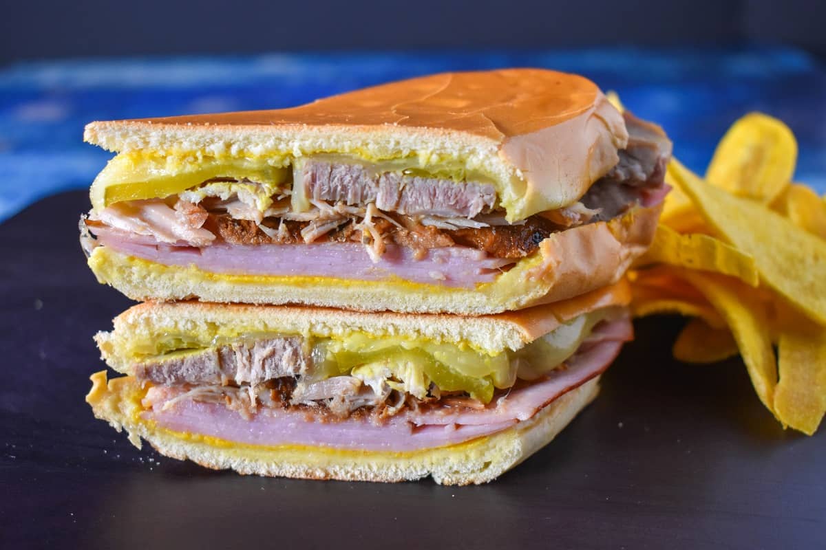 An image of the sandwich one half stacked on the other half with plantain chips on the right side and displayed on a black cutting board on a blue table.