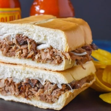 The pan con lechon cut in half and stacked with two bottles of hot sauce and plantain chips in the background.