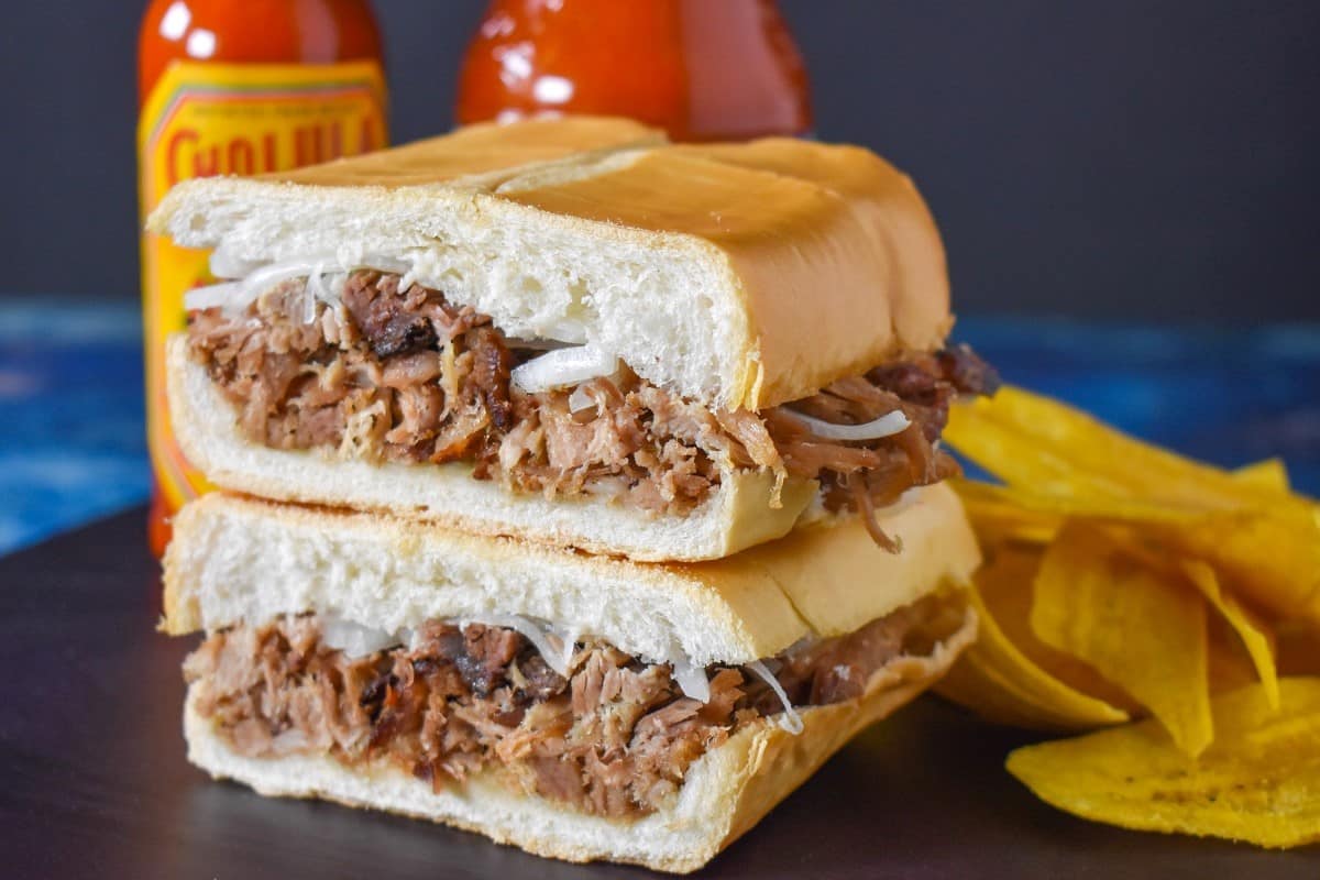 The pan con lechon cut in half and stacked with two bottles of hot sauce and plantain chips in the background.