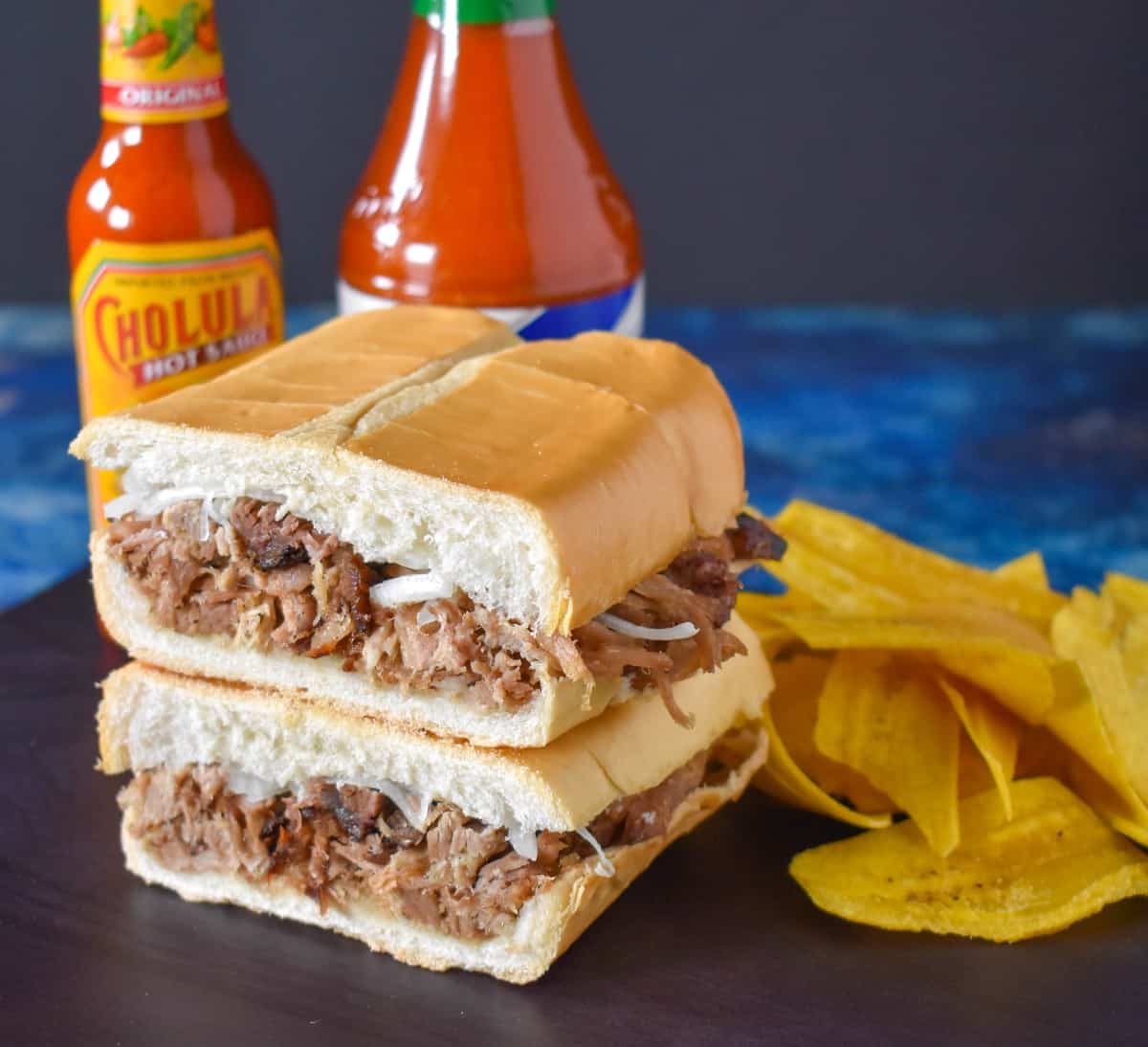 The pan con lechon cut in half and stacked with two bottles of hot sauce and plantain chips in the background, all displayed on a brown cutting board on a blue table.