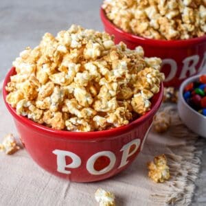 Caramel popcorn piled in a red bowl with more popcorn and colorful candy in the background.