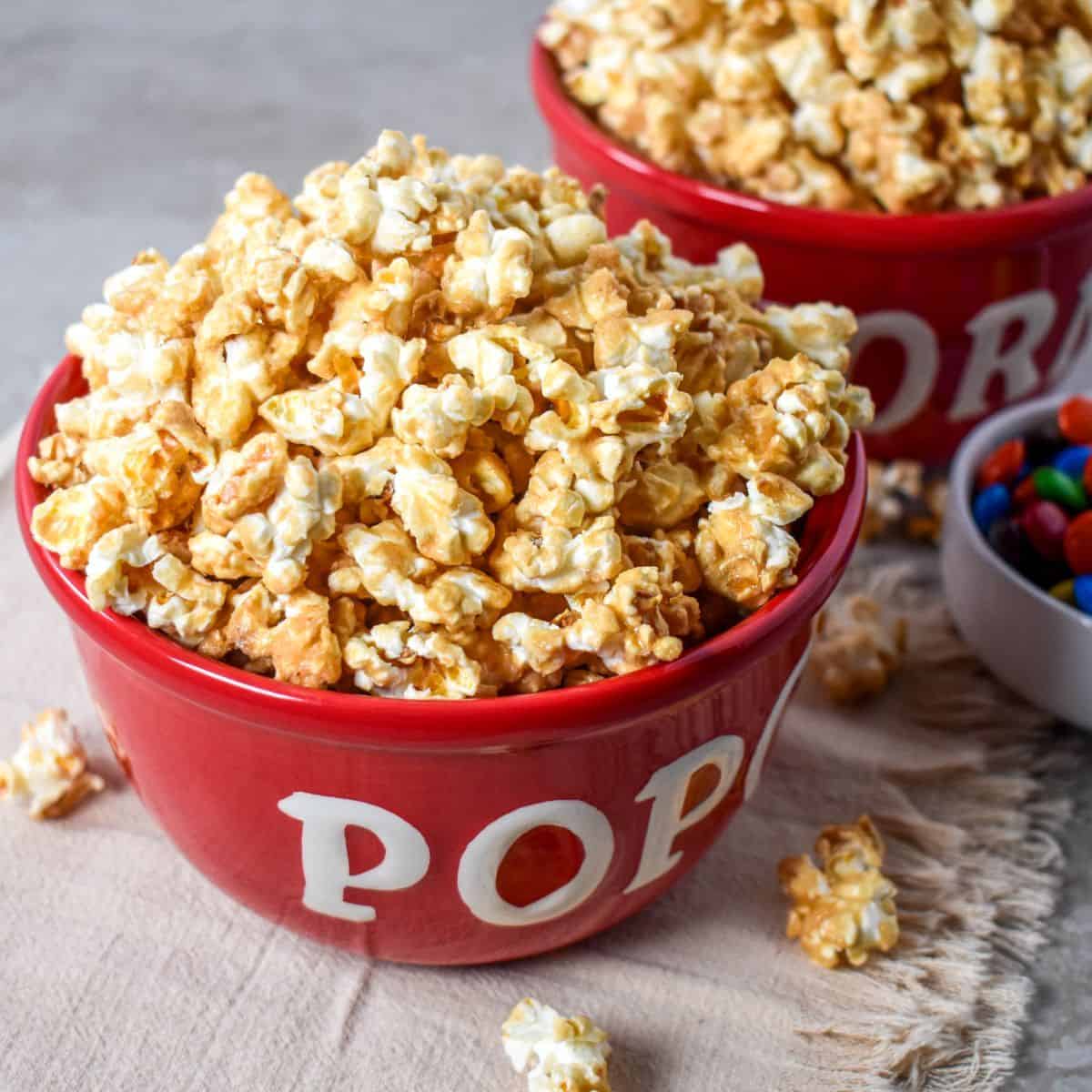 Caramel popcorn piled in a red bowl with more popcorn and colorful candy in the background.
