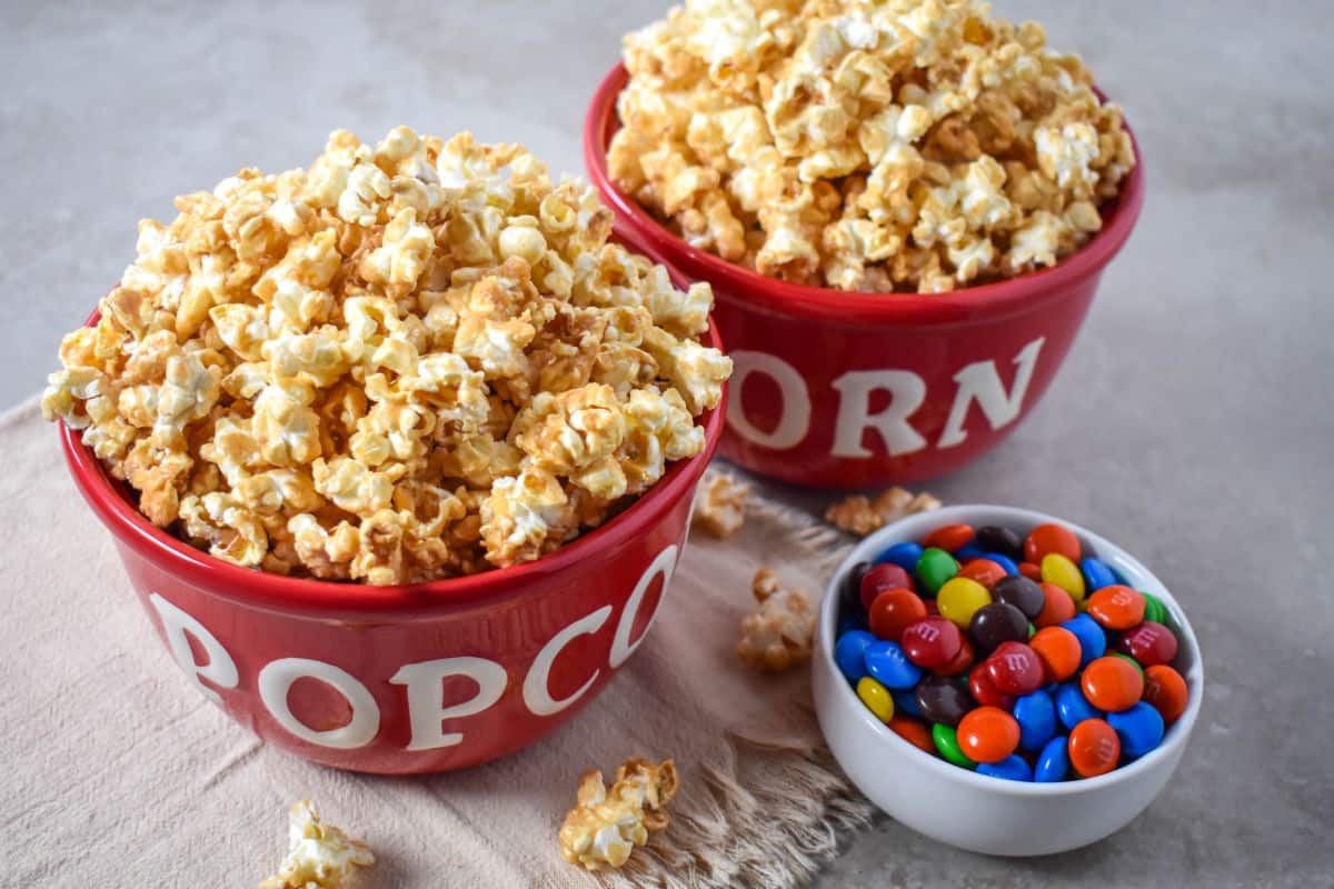 Caramel popcorn in red bowls with colorful candy arranged on a light table.