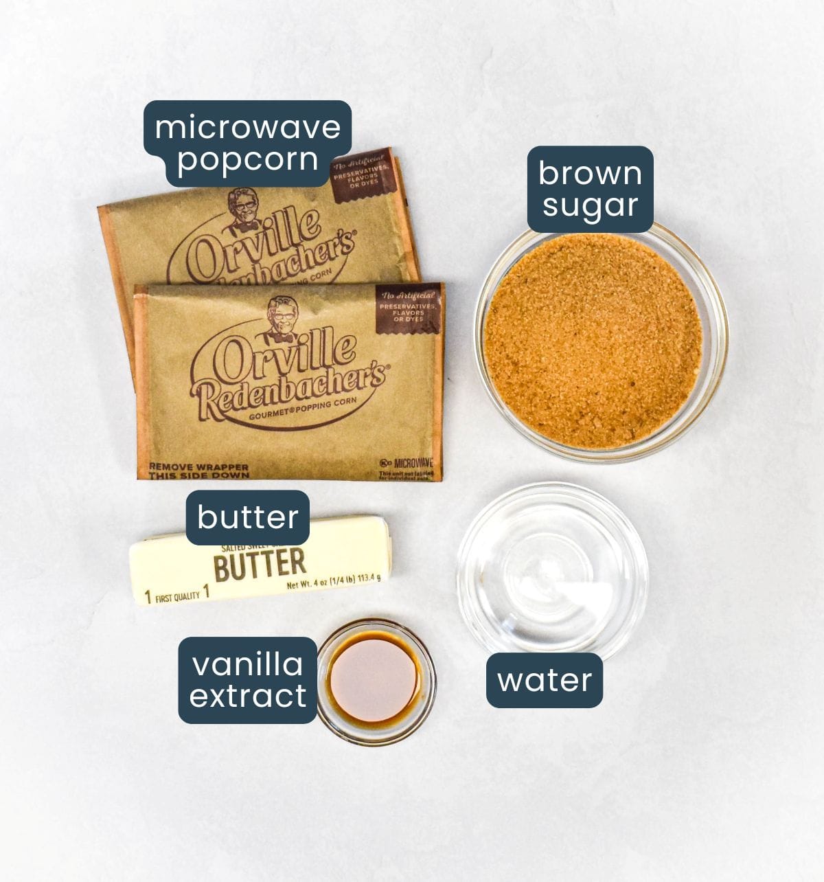 The ingredients for the recipe, microwave popcorn, brown sugar, butter, water, and vanilla extract arranged on a white table, with each one labeled.