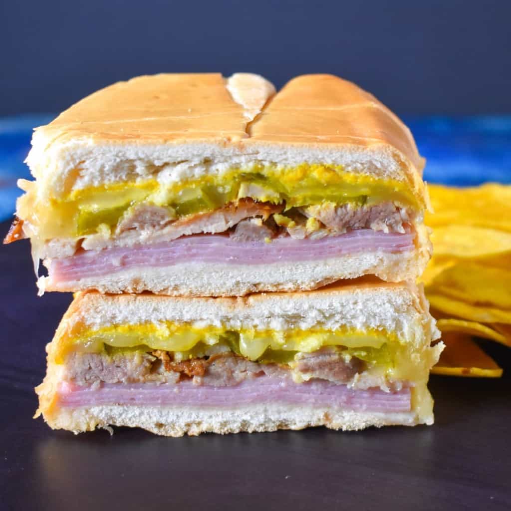 An image of the sandwich one half stacked on the other half with plantain chips on the right side and displayed on a black cutting board on a blue table.