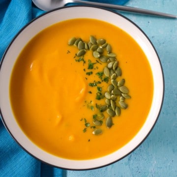 An image of the pumpkin apple soup served in a white bowl with a black rim set on an aqua table with an aqua linen to the left side. The soup is garnished with pumpkin seeds and chopped parsley.