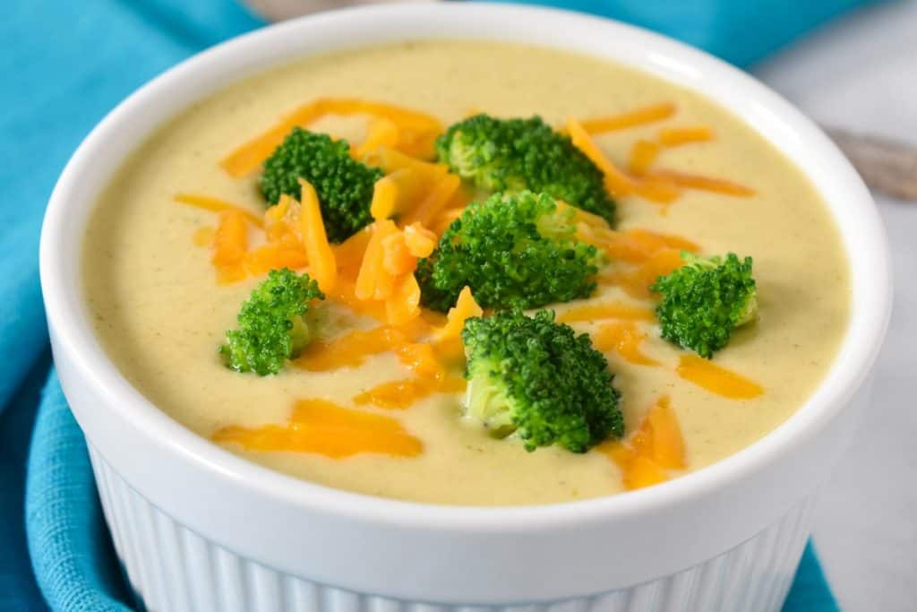 Broccoli cauliflower soup served in a white bowl and garnished with broccoli florets and shredded cheddar cheese.