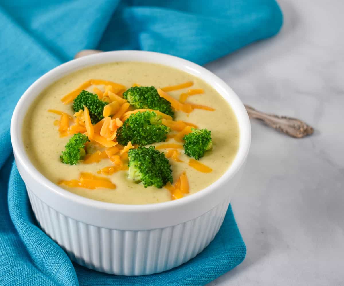 The finished soup served in a white bowl displayed with a teal linen on a white table. The soup is garnished with broccoli florets with shredded cheese.