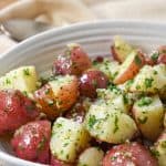 The finished garlic parsley potatoes served in a white bowl on a beige linen with a serving spoon in the background.