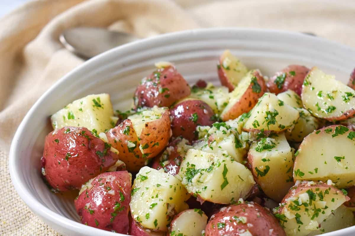 The finished garlic parsley potatoes served in a white bowl on a beige linen with a serving spoon in the background.