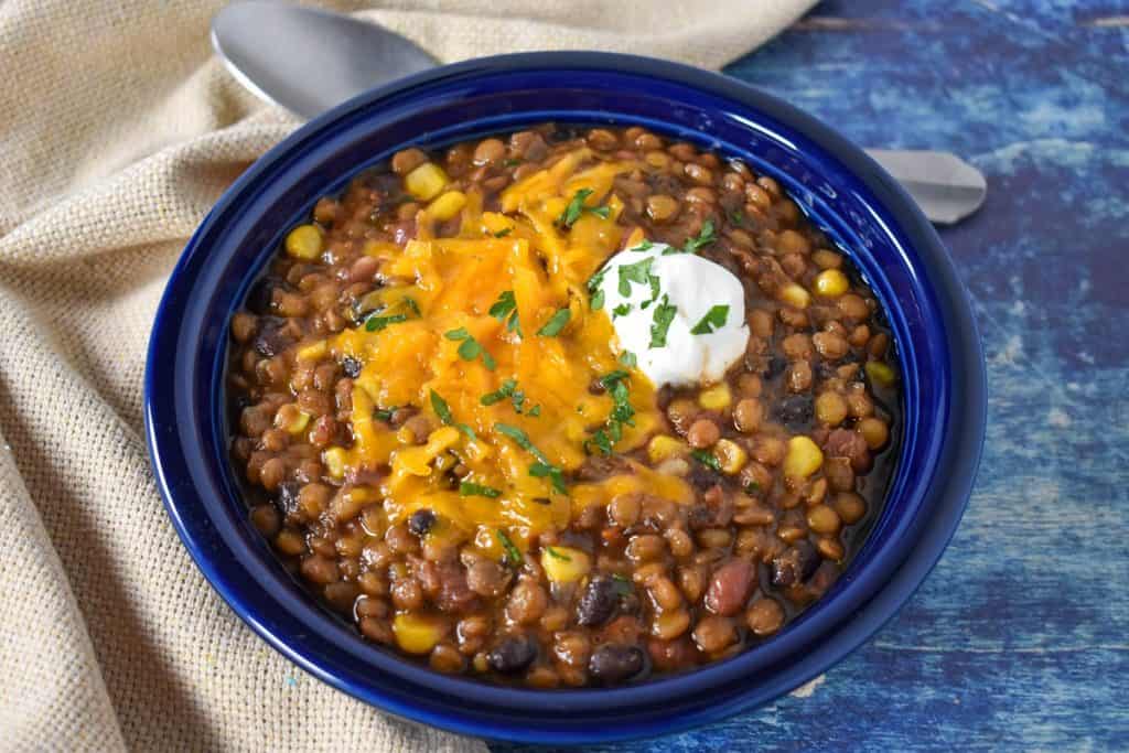 The chili served in a blue bowl and garnished with melted cheddar cheese and a dollop of sour cream displayed on a blue table with a beige linen and a spoon.