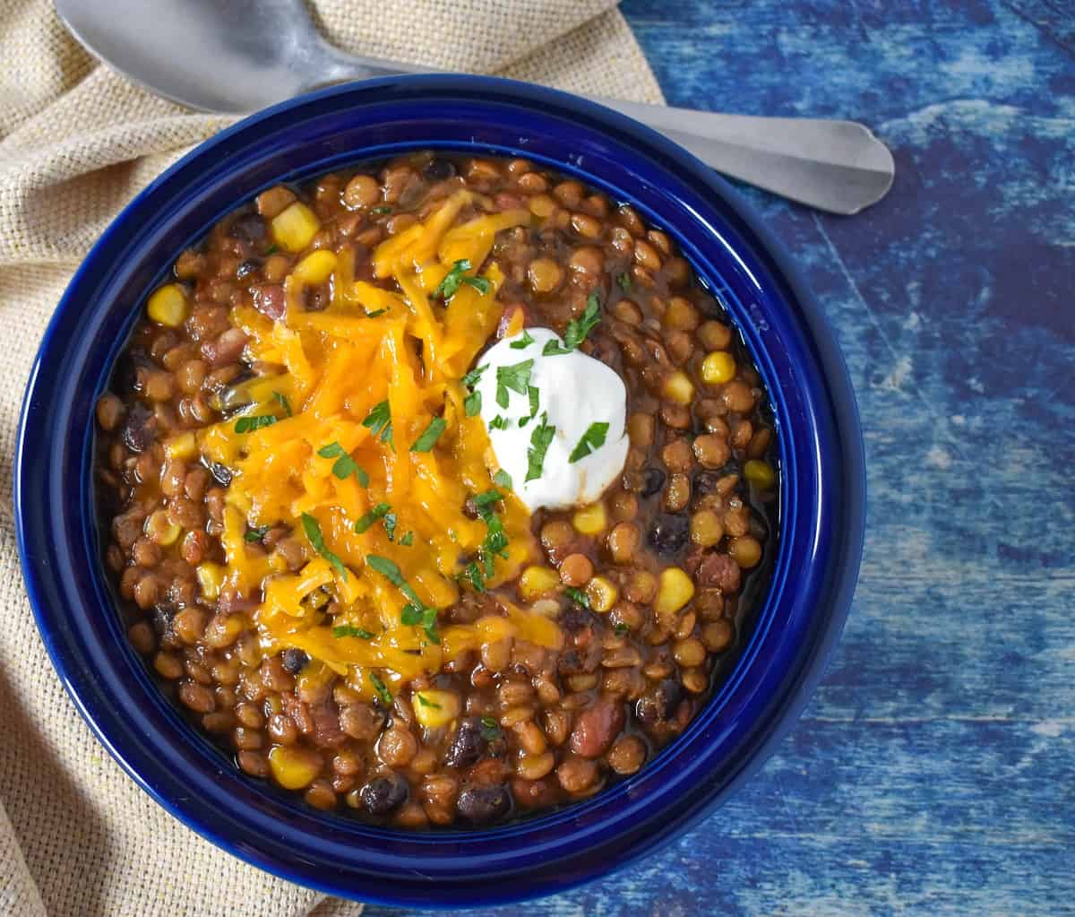 The lentil chili served in a blue bowl with a beige colored linen and a spoon on the side. The chili is garnished with melted cheddar cheese and a dollop of sour cream.