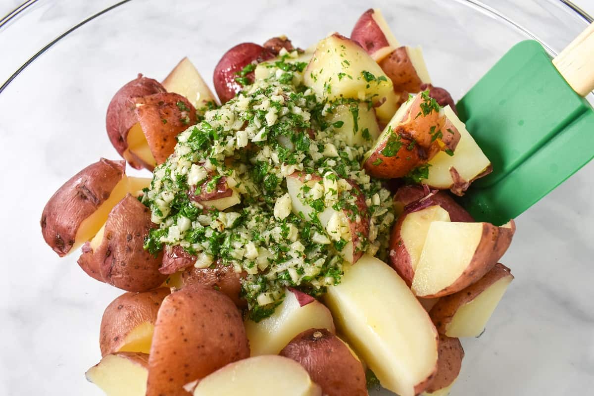 Cut red potatoes in a large, clear bowl with the garlic parsley sauce on top and a green rubber spatula to the right side.